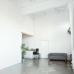 COTAM STUDIO - Jasne, minimalistyczne studio fotograficzne z betonową podłogą, białymi ścianami, dużymi oknami z widokiem na miasto, szarą sofą i aneksem kuchennym z mikrofalówką.