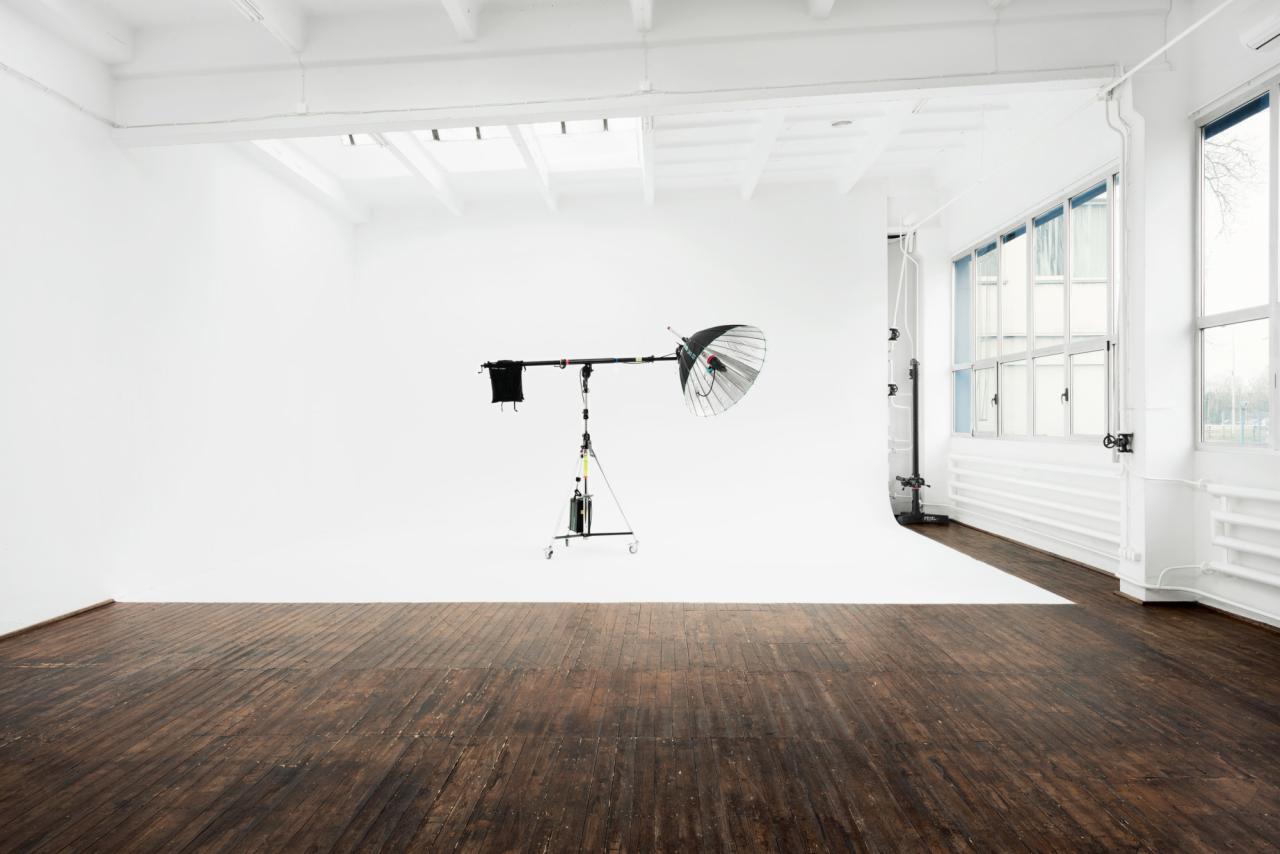 Puste studio fotograficzne z drewnianą podłogą, białym tłem i lampą studyjną z softboxem typu beauty dish, oświetlone naturalnym światłem z okien.