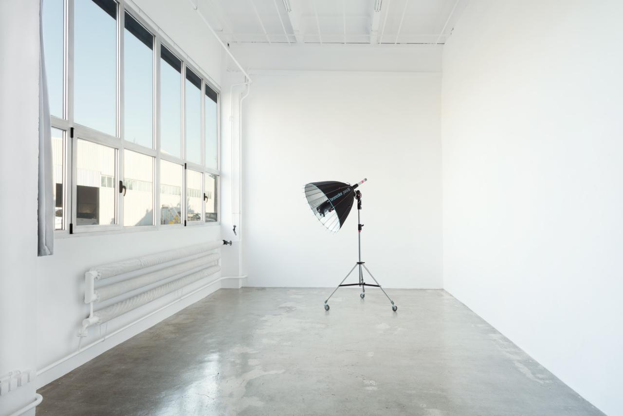 Puste studio fotograficzne z dużym oknem, grzejnikiem owiniętym materiałem ochronnym i lampą błyskową Broncolor na statywie.