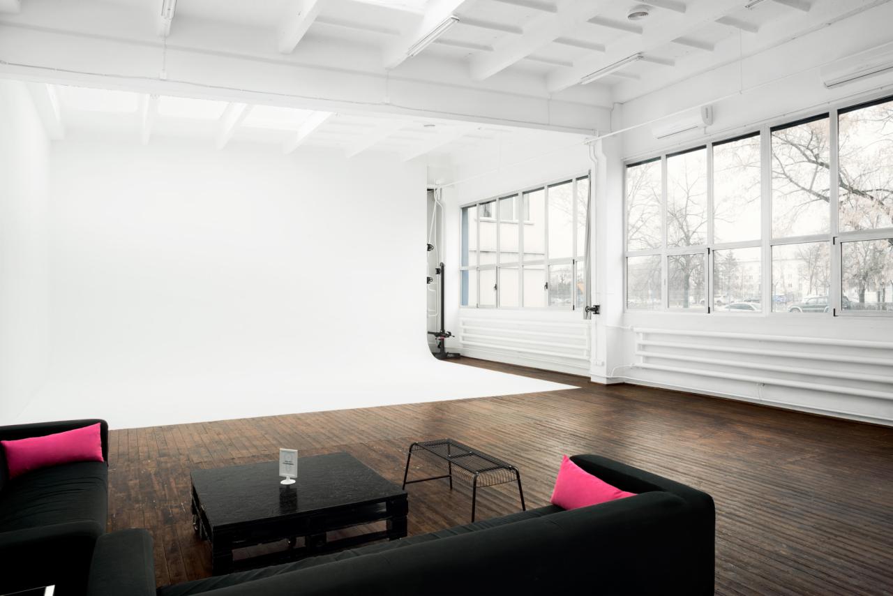 Puste studio fotograficzne z białą cykloramą, dużymi oknami z widokiem na drzewa, drewnianą podłogą i czarną sofą z różowymi poduszkami.