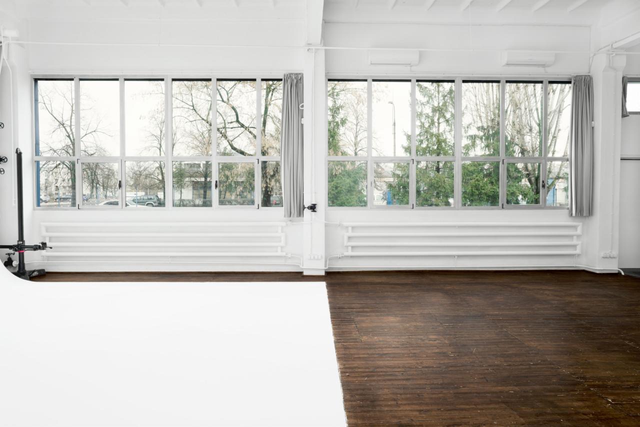 Puste studio fotograficzne z dużymi oknami, białym tłem i drewnianą podłogą, widok na drzewa za oknem.