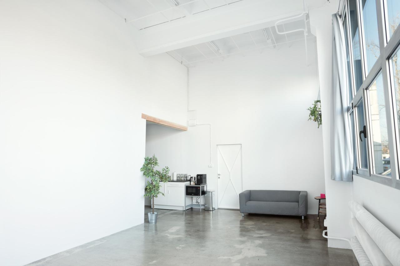 Jasne, minimalistyczne studio fotograficzne z betonową podłogą, białymi ścianami, dużymi oknami z widokiem na miasto, szarą sofą i aneksem kuchennym z mikrofalówką.