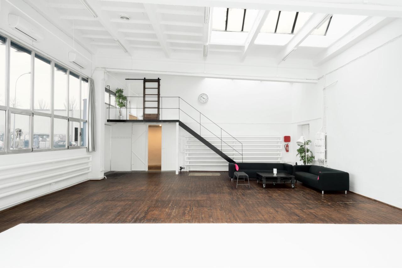 Przestronne, białe studio fotograficzne z drewnianą podłogą, antresolą z drabinką, dużymi oknami i minimalistycznym umeblowaniem: czarne sofy i stolik na kółkach. Widoczne światło dzienne...