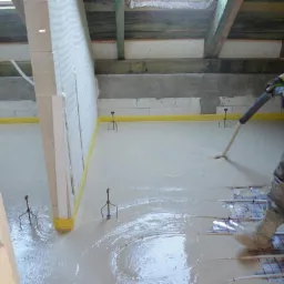 Aplikacja płynnej wylewki na ogrzewanie podłogowe w surowym wnętrzu, widoczne narzędzia i pracownik w odblaskowej kamizelce.