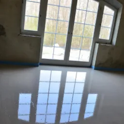 Świeżo wylana, gładka, jasnoszara posadzka z wyraźnym odbiciem okien w nowym domu, zabezpieczona niebieską taśmą przy ścianach z surowym tynkiem.