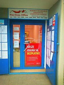 Wejście do biura nieruchomości z niebieskimi drzwiami i tablicą informacyjną po obu stronach. Nad drzwiami szyld z napisem 'Pos-Dom. Office' oraz informacjami o nieruchomościach, kredytach...
