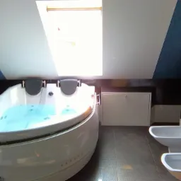 Jasna łazienka na poddaszu ze skośnym sufitem i oknem dachowym, wyposażona w dużą, okrągłą wannę z hydromasażem wypełnioną wodą, bidet i toaletę w nowoczesnym stylu, wykończona ciemnymi płytkami.