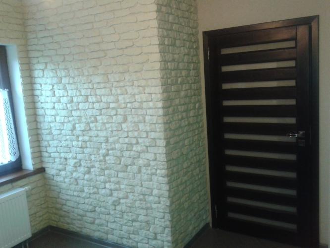 Wykończone wnętrze z dekoracyjną ścianą z imitacji kamienia i nowoczesnymi, ciemnymi drzwiami z poziomymi przeszkleniami. Widoczny fragment okna z firanką i grzejnik.