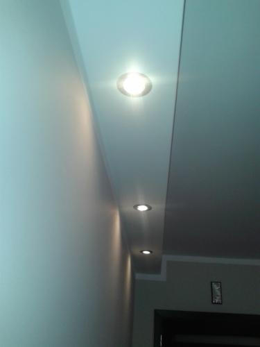 Biały sufit z wbudowanymi, okrągłymi lampami halogenowymi, oświetlającymi pomieszczenie. Widok pod kątem, z fragmentem ściany i drzwi w tle.