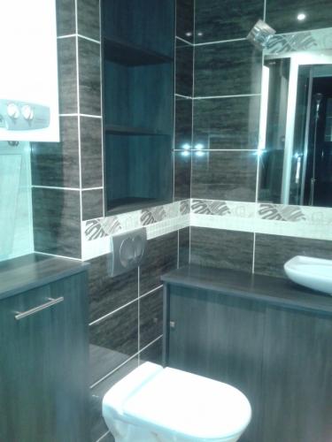 Nowoczesna łazienka z ciemnymi płytkami imitującymi drewno, białym sanitariatem i szafkami pod umywalką. Widoczna wnęka z półkami i podświetlenie.