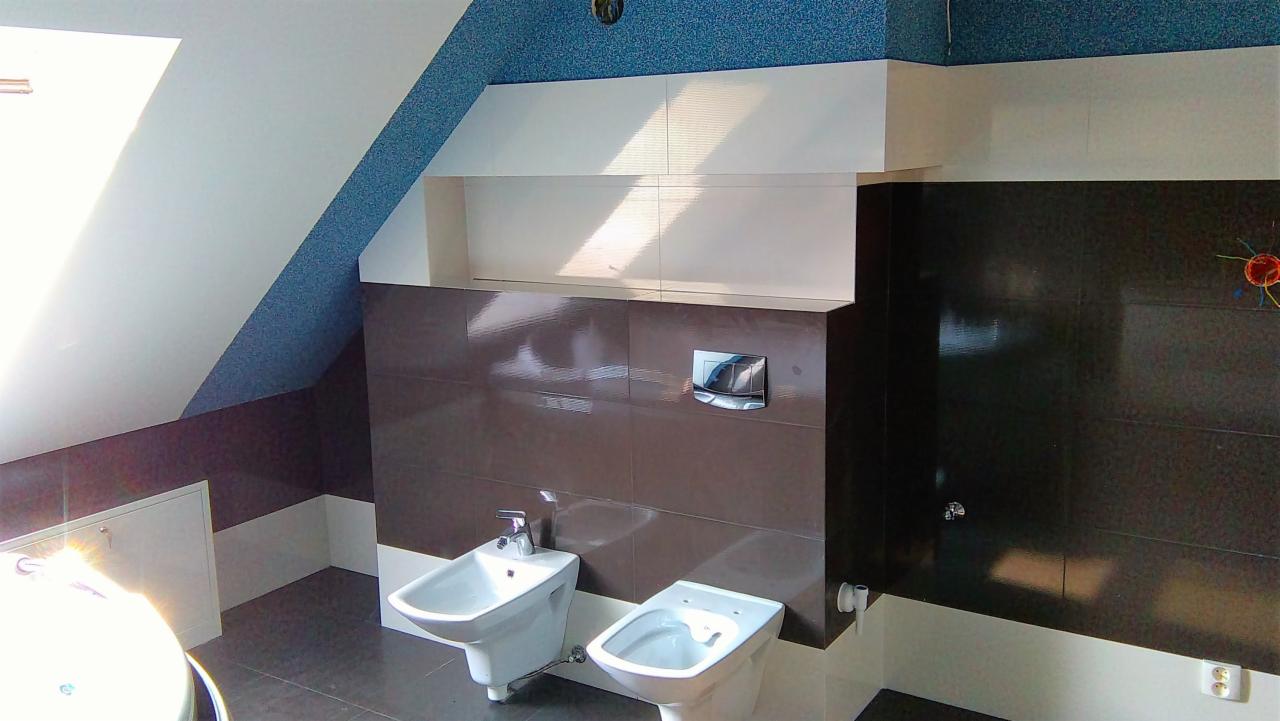 Nowoczesna łazienka na poddaszu z brązowymi i białymi płytkami, wiszącą toaletą i bidetem, oświetlona wpadającym światłem słonecznym przez okno dachowe.