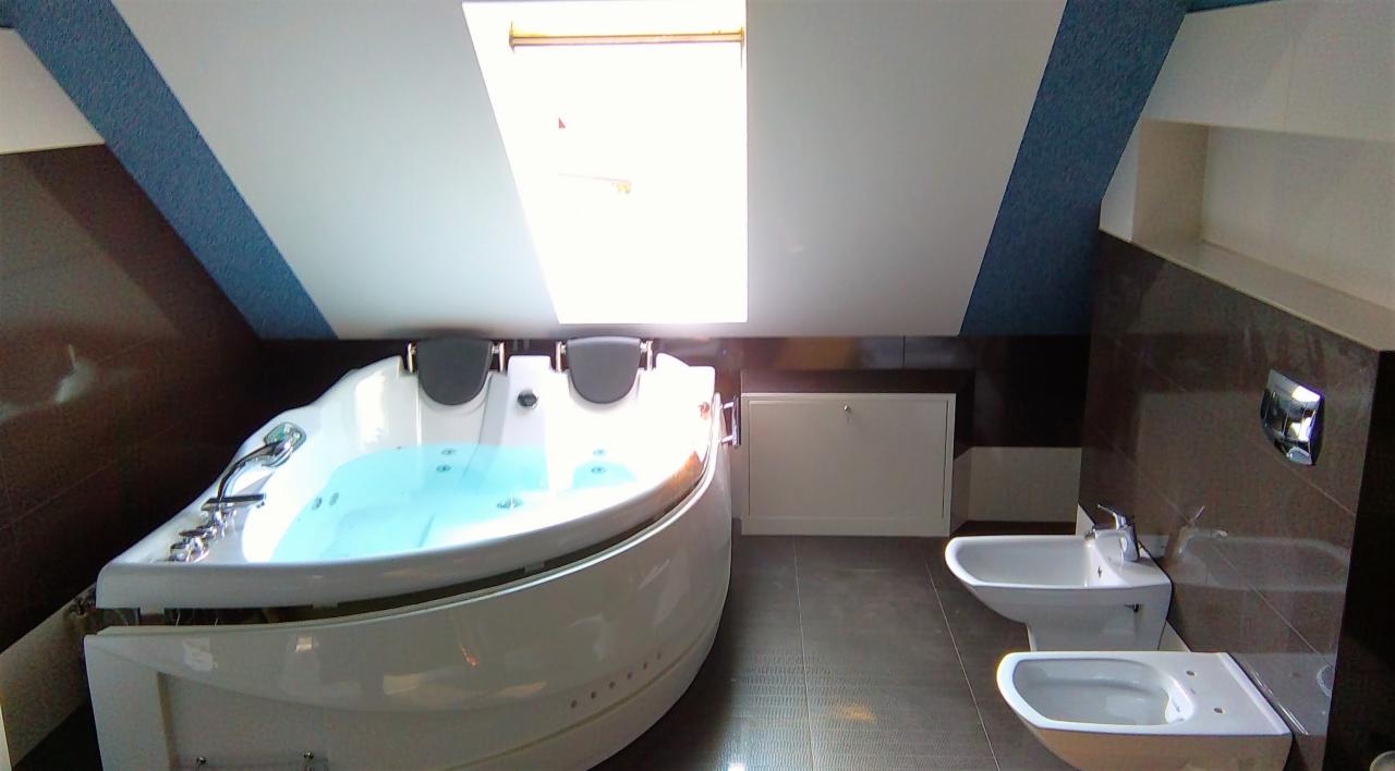 Jasna łazienka na poddaszu ze skośnym sufitem i oknem dachowym, wyposażona w dużą, okrągłą wannę z hydromasażem wypełnioną wodą, bidet i toaletę w nowoczesnym stylu, wykończona ciemnymi płytkami.