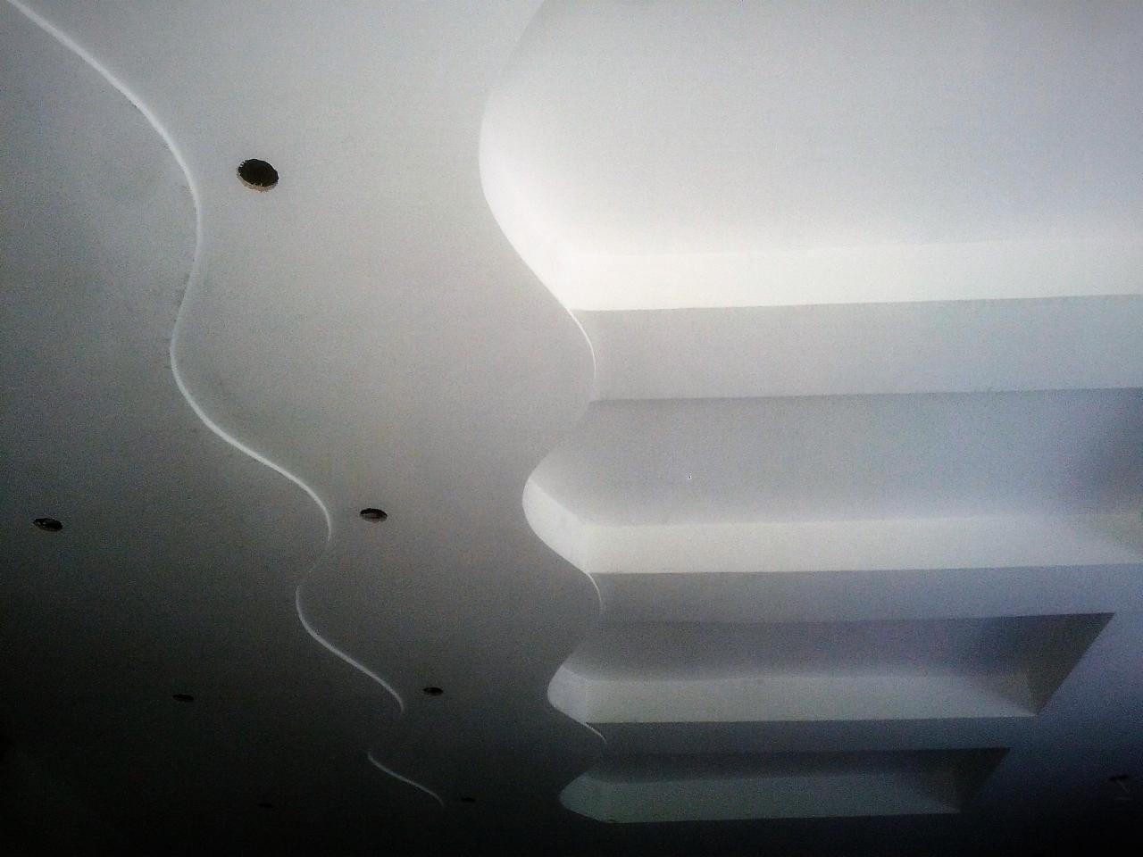 Biały sufit z dekoracyjnymi elementami z płyty gipsowo-kartonowej, z widocznymi otworami na oświetlenie i schodkową konstrukcją po prawej stronie.