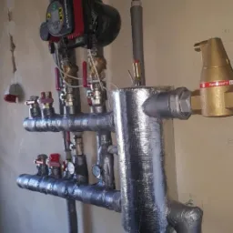 Kompleksowa instalacja grzewcza: pompa obiegowa Grundfos, zbiornik wyrównawczy owinięty folią aluminiową, zawory i manometry na szarych rurach w pomieszczeniu technicznym.