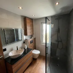 Nowoczesna łazienka z drewnianymi elementami, marmurową umywalką i kabiną prysznicową typu walk-in z czarnym wykończeniem, oświetlona punktowymi lampami sufitowymi.