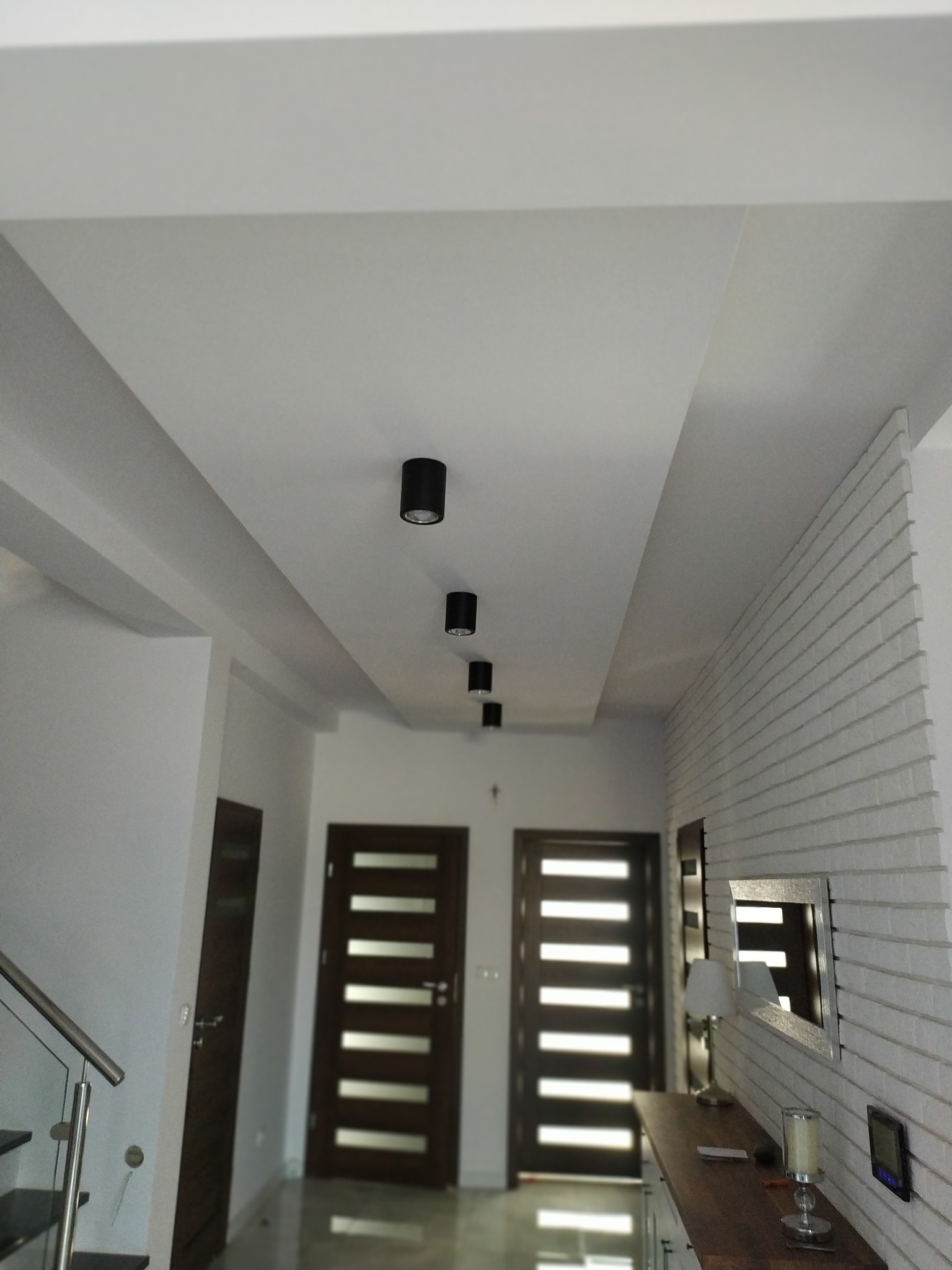 Nowoczesny przedpokój z podwieszonym sufitem, oświetleniem punktowym, ścianą z białej cegły i dwiema parami drzwi z przeszkleniami.