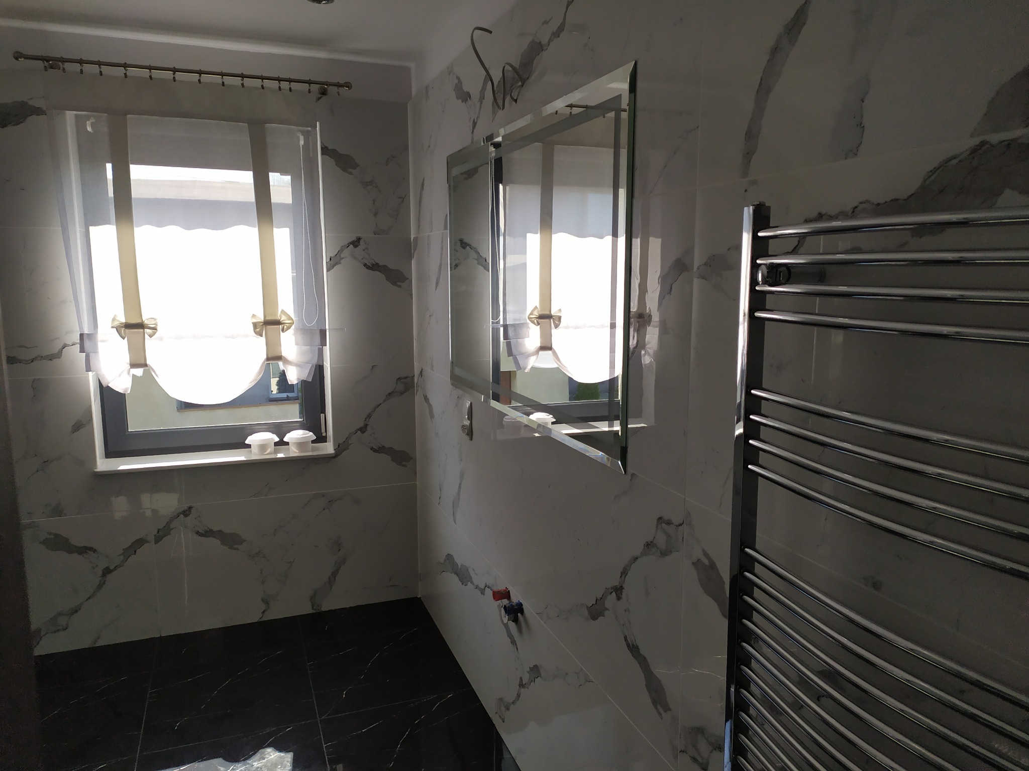 Nowoczesna łazienka z marmurowymi płytkami na ścianach, czarną podłogą, grzejnikiem drabinkowym i lustrem bez ramy. W oknie widoczne białe rolety rzymskie.