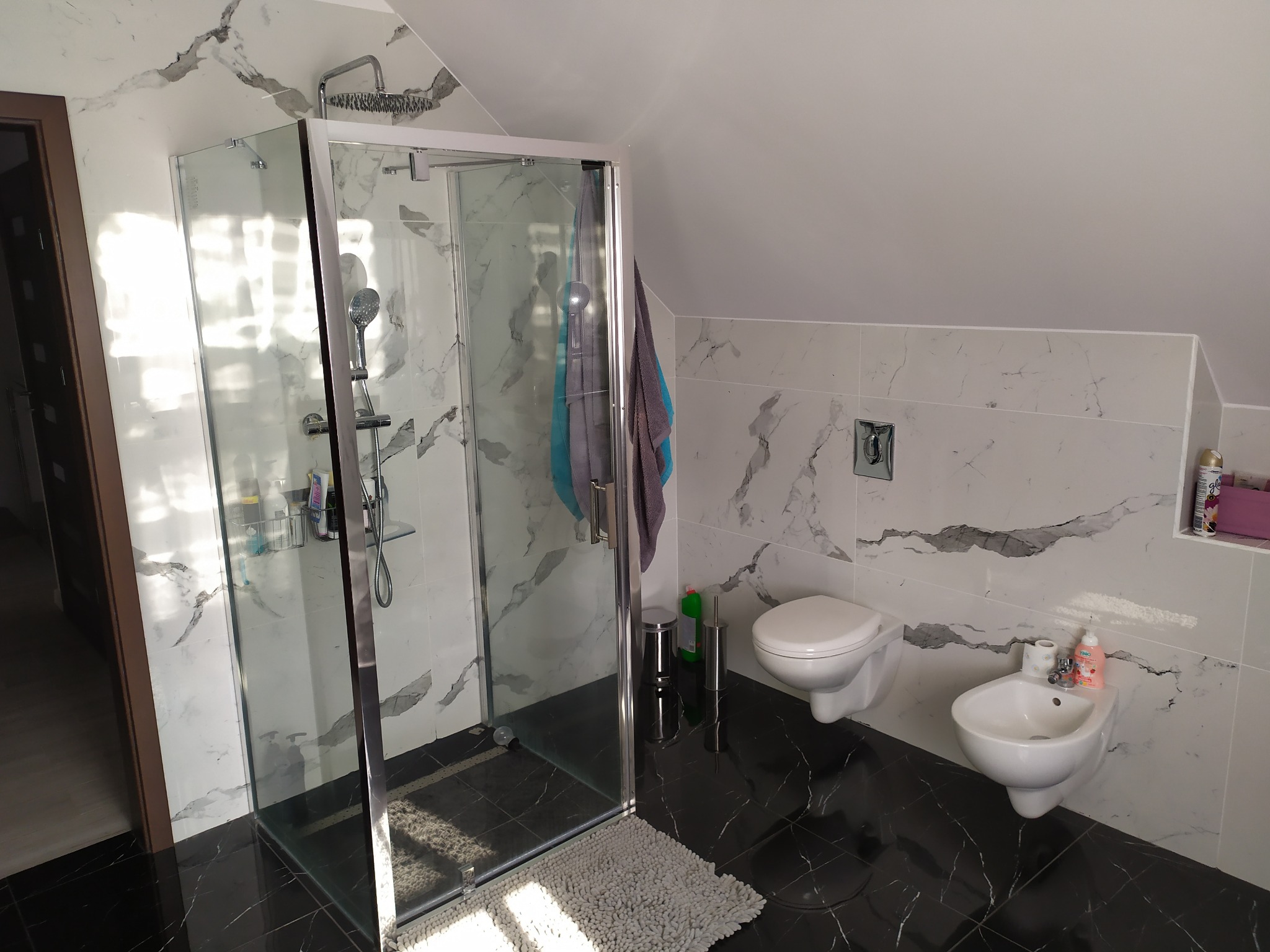 Nowoczesna łazienka na poddaszu z czarno-białymi marmurkowymi płytkami, kabiną prysznicową ze srebrnymi ramami, wiszącą toaletą i bidetem oraz szarym dywanikiem przed kabiną.