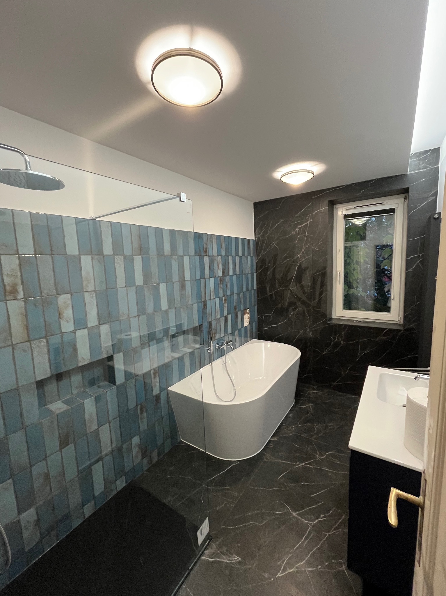 Nowoczesna łazienka z niebieskimi, pionowymi płytkami na ścianie prysznicowej, wolnostojącą białą wanną, czarną marmurową podłogą i ścianą, oraz oknem z widokiem na zieleń.