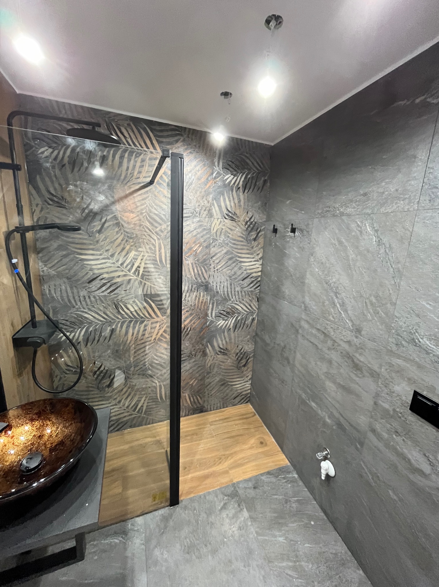 Nowoczesna łazienka z kabiną prysznicową typu walk-in, umywalką nablatową w odcieniach brązu i szarości oraz płytkami ściennymi z motywem liści palmowych i imitacji kamienia.
