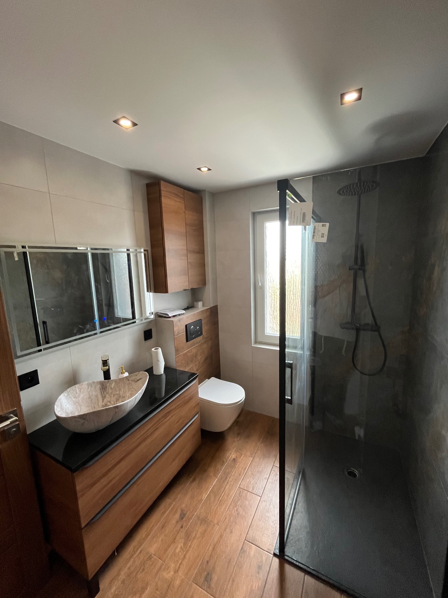 Nowoczesna łazienka z drewnianymi elementami, marmurową umywalką i kabiną prysznicową typu walk-in z czarnym wykończeniem, oświetlona punktowymi lampami sufitowymi.