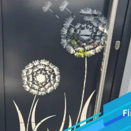 Nowoczesna, grafitowa brama wejściowa z dekoracyjnym motywem dmuchawca i srebrnym uchwytem, widziana pod kątem.