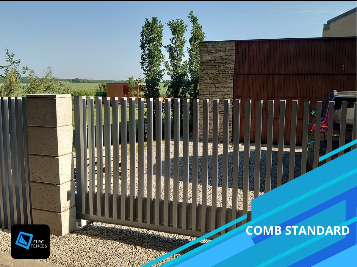 Nowoczesna brama wjazdowa, pionowe profile w kolorze szarym, słupki betonowe, żwirowe podłoże, w tle dom z elementami drewna i kamienia.