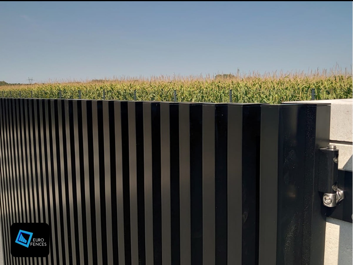 Nowoczesna, czarna brama z pionowych profili metalowych na betonowym słupku, z widocznymi zawiasami, w tle pole kukurydzy pod błękitnym niebem.