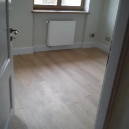 Jasny, drewniany parkiet w pokoju z białymi listwami przypodłogowymi i grzejnikiem pod oknem, widok zza uchylonych, białych drzwi.