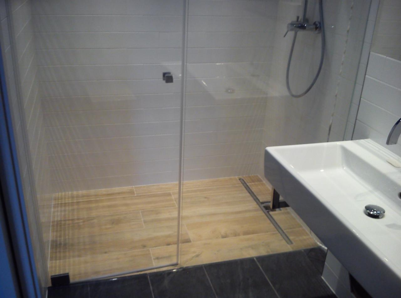 Nowoczesna kabina prysznicowa z drewnopodobną podłogą i minimalistyczną umywalką. Białe, poziome płytki ścienne. Widoczny odpływ liniowy. Styl skandynawski.