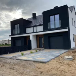 Nowoczesny dwukondygnacyjny dom bliźniak z czarno-białą elewacją i szarym brukowanym podjazdem, widok z poziomu gruntu, pochmurne niebo w tle.