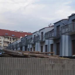 Szereg nowych budynków mieszkalnych z balkonami, świeżo po malowaniu elewacji na szaro, z stertą drewna i zabezpieczeniami budowlanymi na pierwszym planie.