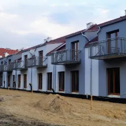 Szereg nowych domów z balkonami i elewacją w odcieniach szarości, teren przed budynkami w trakcie prac ziemnych.
