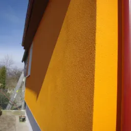 Żółta fasada domu z widoczną rurą spustową w kolorze czerwonym, drabina oparta o ścianę. Świeżo pomalowana elewacja w słoneczny dzień.