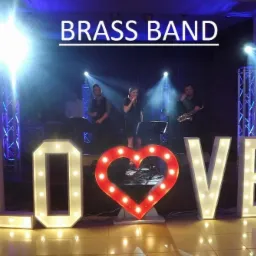 Zespół na scenie za podświetlanym napisem LOVE z żarówkami, saksofonista po prawej, wokalistka pośrodku, oświetlenie sceniczne.