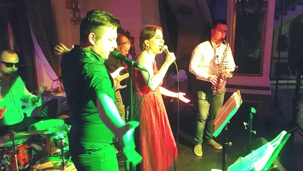 Zespół muzyczny podczas występu na scenie: wokalistka w czerwonej sukni, saksofonista, gitarzysta i perkusista w akcji.