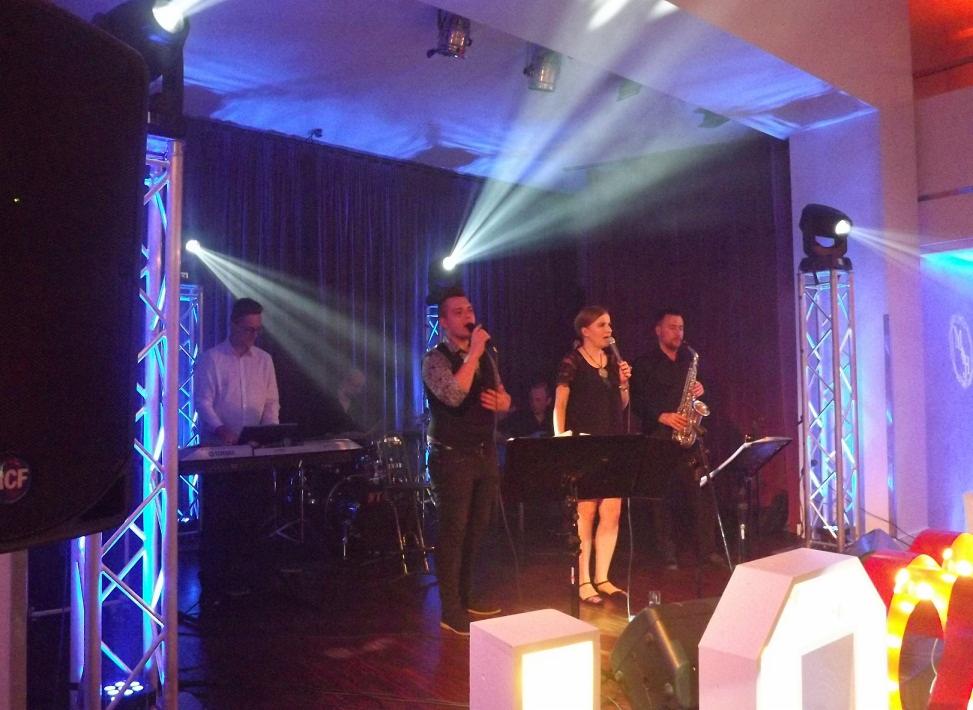 Zespół muzyczny na scenie podczas występu, z oświetleniem punktowym i dekoracjami świetlnymi na podłodze.