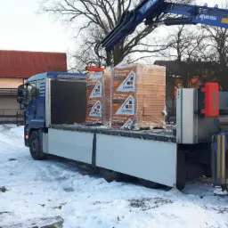 Ciężarówka z dźwigiem Atlas i paletami z materiałem budowlanym Ceramiczna perfekcja na zaśnieżonym placu, w tle budynki mieszkalne.