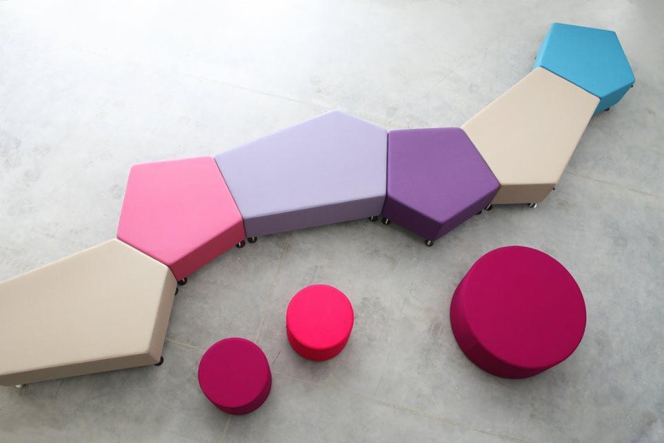 Modułowy zestaw siedzisk w pastelowych kolorach: różowy, fioletowy, beżowy i niebieski, ułożony w zygzak na jasnej podłodze, uzupełniony o okrągłe pufy w odcieniach różu.