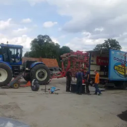 Wulkanizacja mobilna przy niebieskim ciągniku New Holland T9060 z podpiętym czerwonym agregatem uprawowym, grupa mechaników pracuje nad oponami, w tle budynek z czerwonej cegły i drzewa.