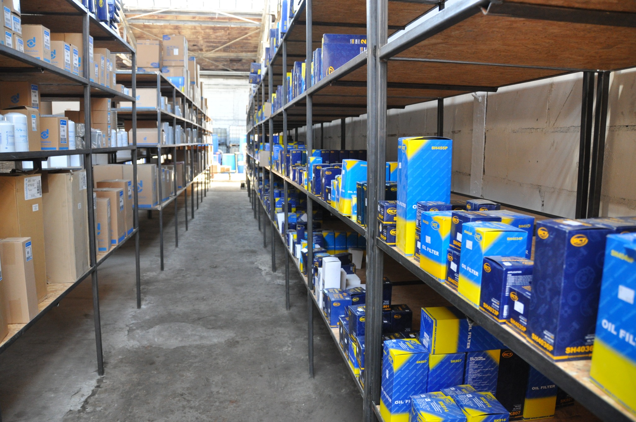 Magazyn z metalowymi regałami wypełnionymi kartonami z filtrami oleju samochodowego, widok w głąb alejki między regałami, betonowa podłoga.