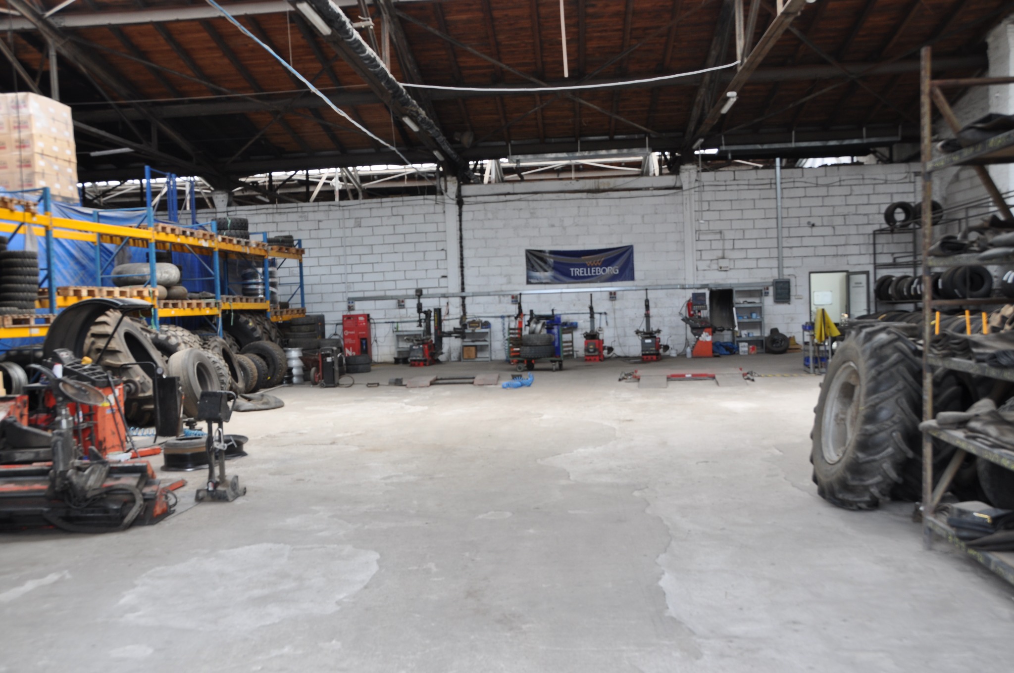 Wnętrze warsztatu z oponami różnej wielkości, narzędziami i sprzętem do naprawy opon. W tle widoczny baner reklamowy firmy Trelleborg.