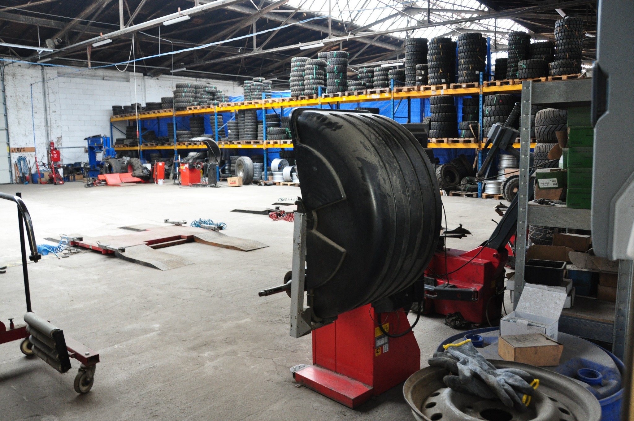 Wnętrze warsztatu samochodowego z maszyną do wyważania kół na pierwszym planie, w tle regały z oponami i podnośnik samochodowy.