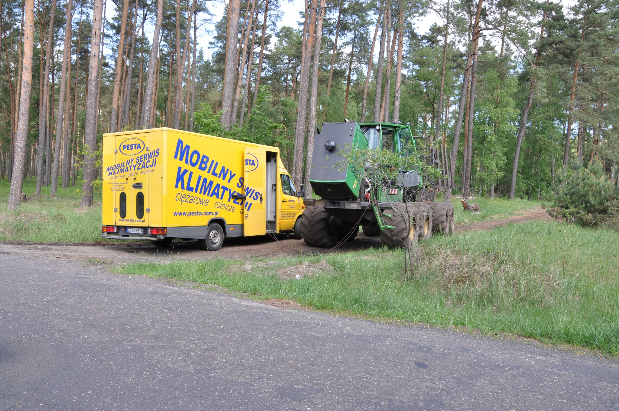 Żółty samochód dostawczy z napisem 'Mobilny Serwis Klimatyzacji' marki Pesta, obsługujący ciężarowe, rolnicze i osobowe pojazdy, zaparkowany obok zielonego kombajnu leśnego w pobliżu lasu.