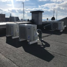 EKO-KLIMA - Trzy jednostki zewnętrzne klimatyzacji marki AUX zainstalowane na płaskim dachu budynku z widokiem na miasto i kościół w oddali, przy słonecznej pogodzie.
