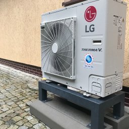 EKO-KLIMA - Zewnętrzna jednostka pompy ciepła LG Therma V, zamontowana na grafitowym stelażu na kostce brukowej, widoczne kropelki wody na obudowie, budynek w tle.