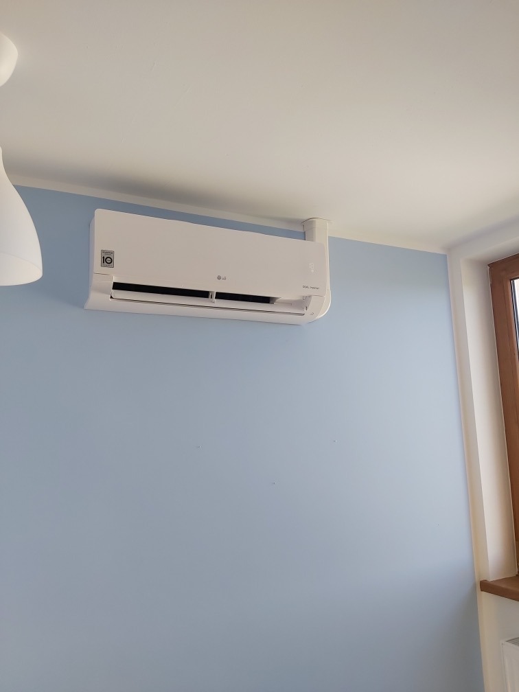 Biała jednostka klimatyzacyjna LG zamontowana na jasnoniebieskiej ścianie w pokoju z widocznym fragmentem okna i lampy sufitowej.