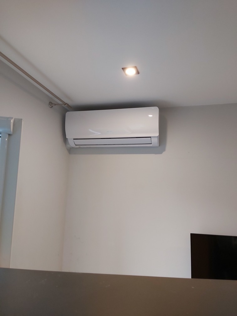 Biała jednostka klimatyzacyjna zamontowana na ścianie w jasnym pomieszczeniu, widoczna z dołu, z elementami instalacji na suficie.