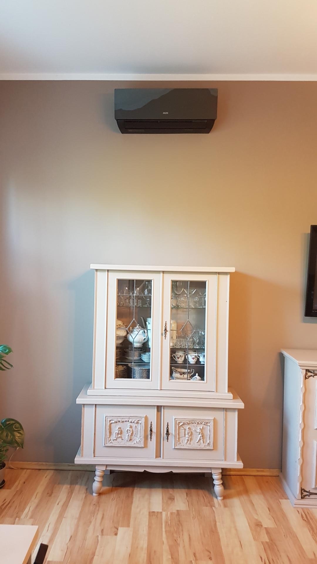Czarny klimatyzator ścienny marki AUX zamontowany nad stylizowaną, kremową witryną w salonie z drewnianą podłogą.