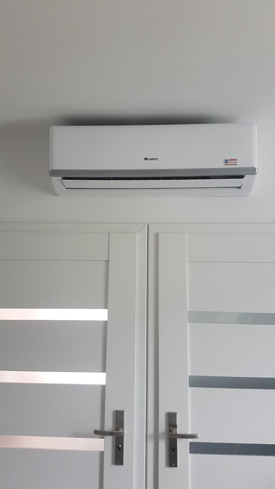 Biała jednostka klimatyzacyjna marki Gree zamontowana na ścianie nad białymi drzwiami z poziomymi, szklanymi paskami.
